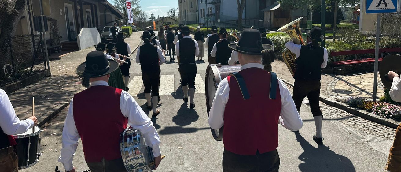 Eine Blaskapelle in traditioneller bayerischer Kleidung geht eine Dorfstraße entlang. Sie tragen verschiedene Instrumente, darunter Trommeln und Blechblasinstrumente.