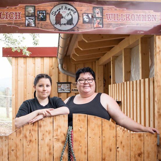 Zwei Frauen stehen vor einer Holzstruktur mit einem Schild, das 'Sweet Home Alpaches' steht. Sie posieren für ein Foto vor einem Tor.