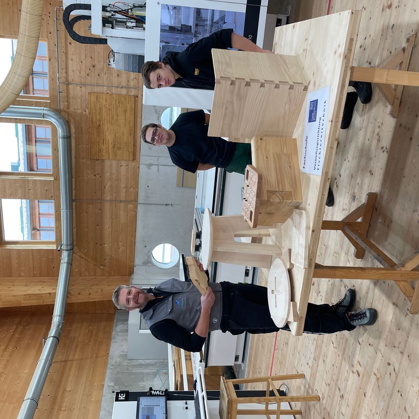 Drei Männer in einer Werkstatt mit Holztischen und Bänken. Zwei halten Holzstücke in den Händen und einer steht und lächelt. Ein Whiteboard mit Text steht auf dem Tisch.