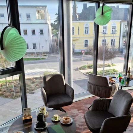 Ein Raum mit großen Fenstern mit Blick auf eine Straße. Ein Tisch hat Essen, Getränke und Bücher. Stühle sind im Raum platziert, mit Blick auf Gebäude draußen.