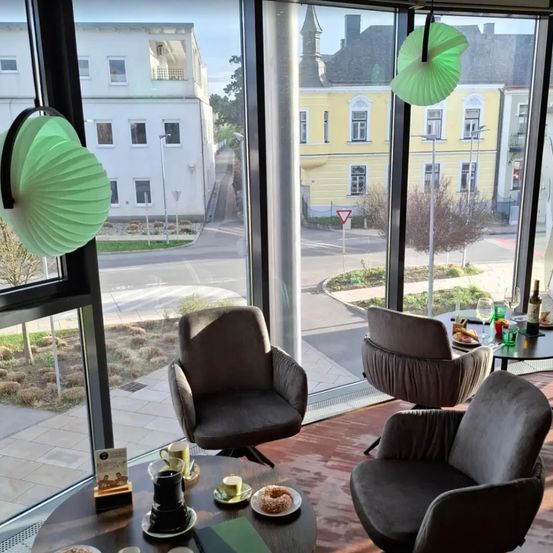 Ein Raum mit großen Fenstern mit Blick auf eine Straße. Ein Tisch hat Essen, Getränke und Bücher. Stühle sind im Raum platziert, mit Blick auf Gebäude draußen.