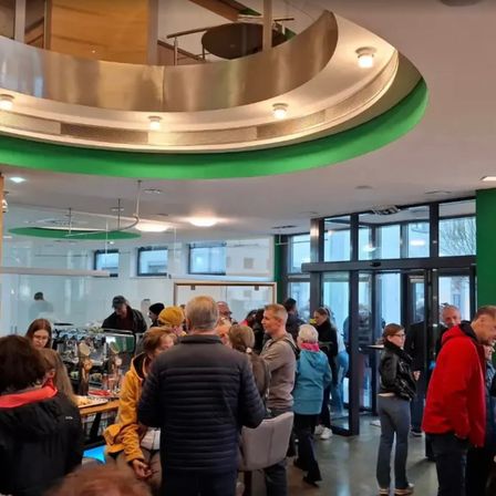 Eine Gruppe von Menschen versammelt sich in einem modernen Gebäude, wahrscheinlich einem Büro. Sie stehen und unterhalten sich. Einige sind in der Nähe eines Tresens, andere um einen Tisch mit einer Kaffeemaschine. Die Decke ist hoch mit metallischen und gläsernen Elementen.