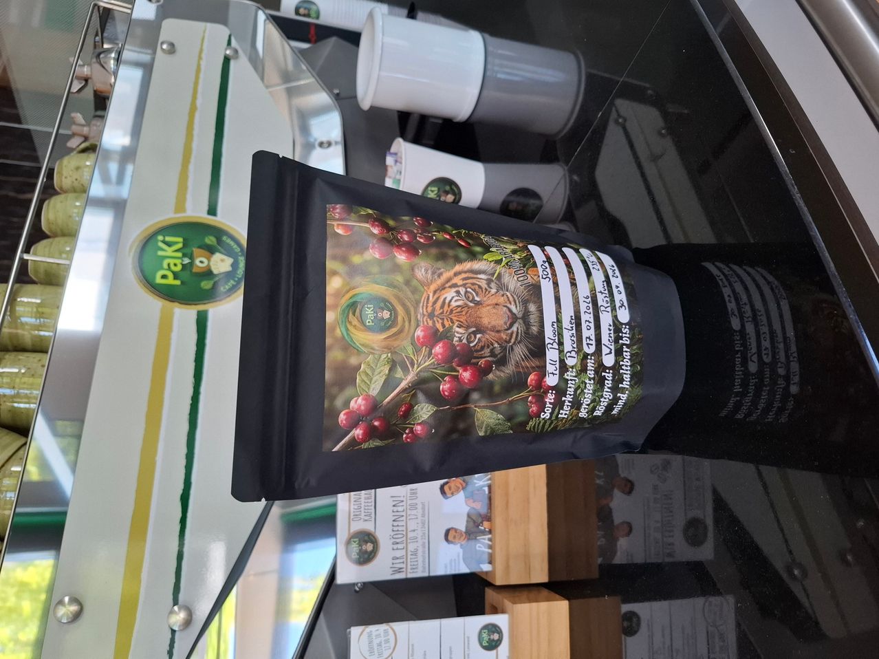 Ein schwarzer Beutel mit einem Tiger und Kaffeebohnen darauf, ausgestellt auf einem Glasregal mit anderen Produkten im Hintergrund.