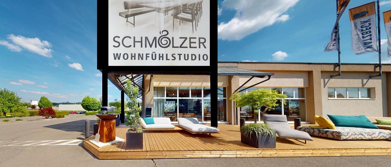 Das Äußere des Schmolzer Living Studios zeigt ein großes Schild, eine Holzterrasse mit Liegestühlen und Topfpflanzen. Das Studio hat Glasfenster und einen Parkplatz.