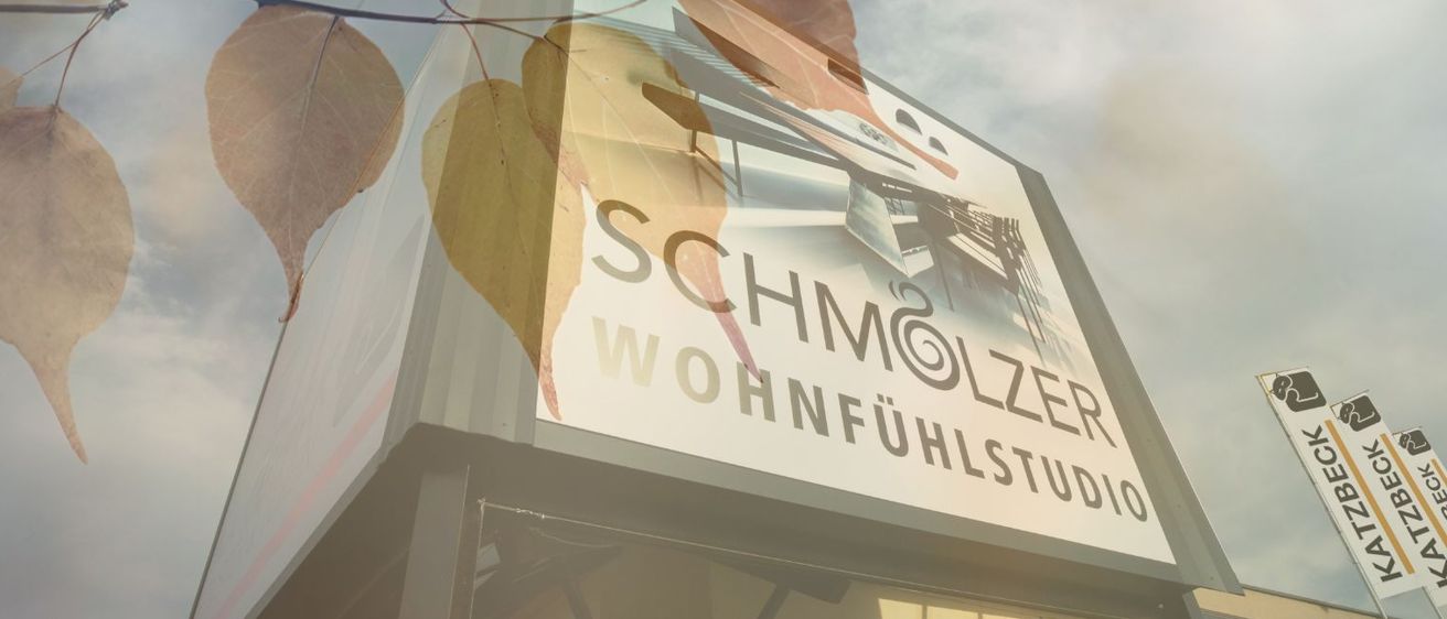 Ein Werbeschild für 'SCHMÖLZER WOHNFÜHLSTUDIO' zeigt ein Bild eines Gebäudes mit Herbstblättern im Vordergrund.
