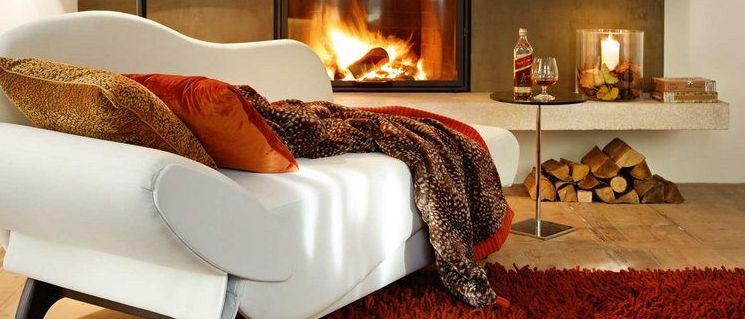 Ein modernes weißes Lounge-Sofa in der Nähe eines Kamins mit einer brennenden Kerze und einem Glas Whiskey auf einem Tisch.