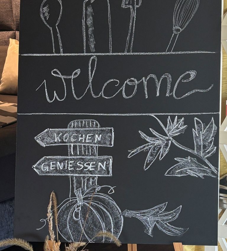 Ein Kreidetafel zeigt das Wort 'Willkommen' mit einem Kürbis und Blättern. Zwei Pfeile zeigen auf 'Kochen' und 'Geniessen' und weisen auf die Richtungen zum Kochen und Genießen hin.