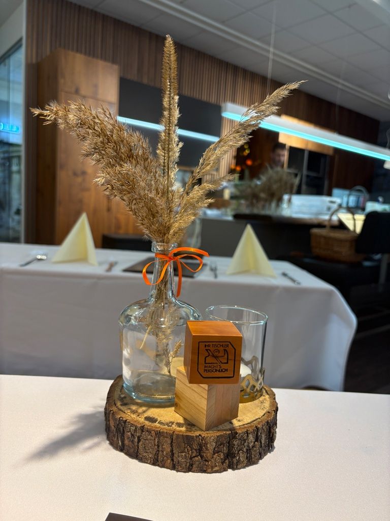 Ein Tischsetting mit einer Vase mit getrockneten Blumen, einem Holzkasten und einem Glas, in einem Raum mit einer Holzwand und einer beleuchteten Deckenlampe.