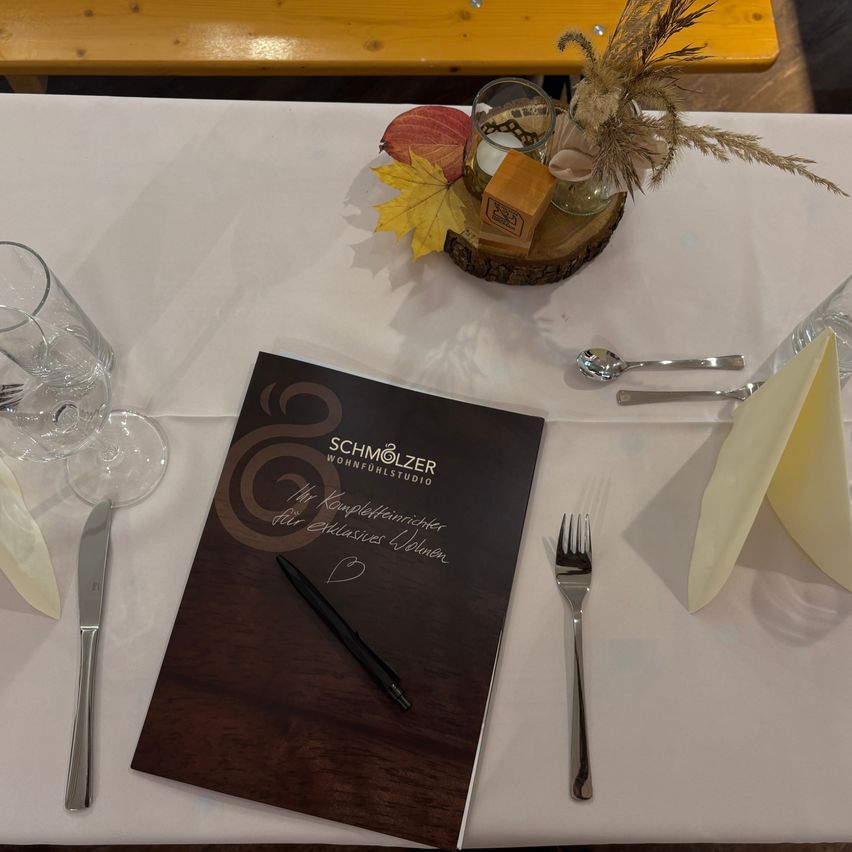 Ein Tisch für ein Esserlebnis mit Besteck, Menü und einer dekorativen Tischmitte. Gläser, ein Löffel und eine Gabel sind arrangiert. Das Menü lautet 'SCHMÖLZER WOHNFLEHSTUDIO'.