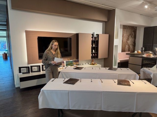 Eine Frau steht in einem Raum mit zwei Tischen, die mit weißen Tüchern bedeckt sind, und hält ein Papier in der Hand, während ein Fernseher und Regale im Hintergrund zu sehen sind.