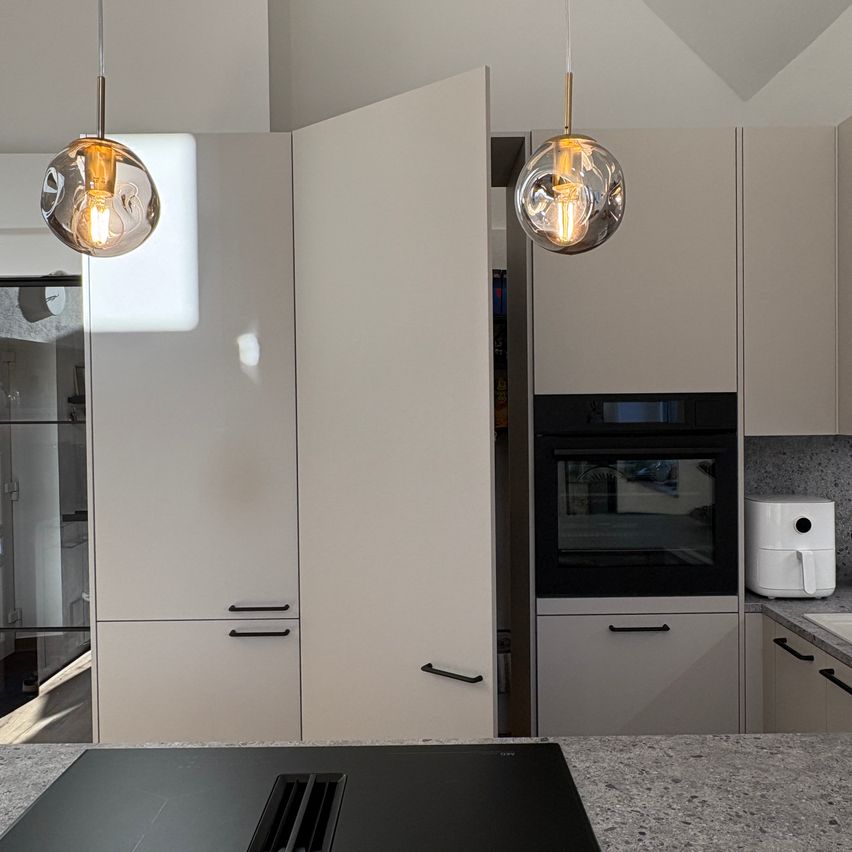 Eine moderne Küche mit weißen Schränken, einer offenen Tür, die einen Einbauofen zeigt, und zwei hängenden Lampen, die den Raum beleuchten.
