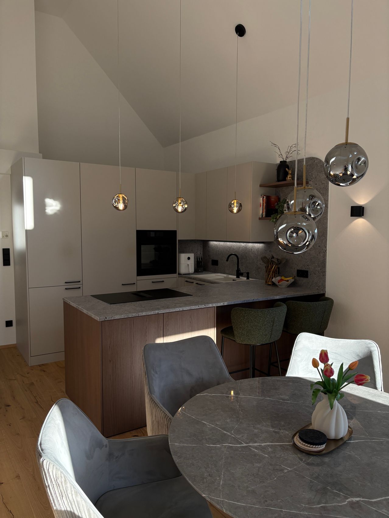 Eine moderne Küche mit weißen Schränken, einer grauen Arbeitsplatte, einem schwarzen Herd und hängenden Lampen. Die Küche verfügt über ein Waschbecken, einen Wasserhahn, einen Holzboden und einen Esstisch mit Blumen.