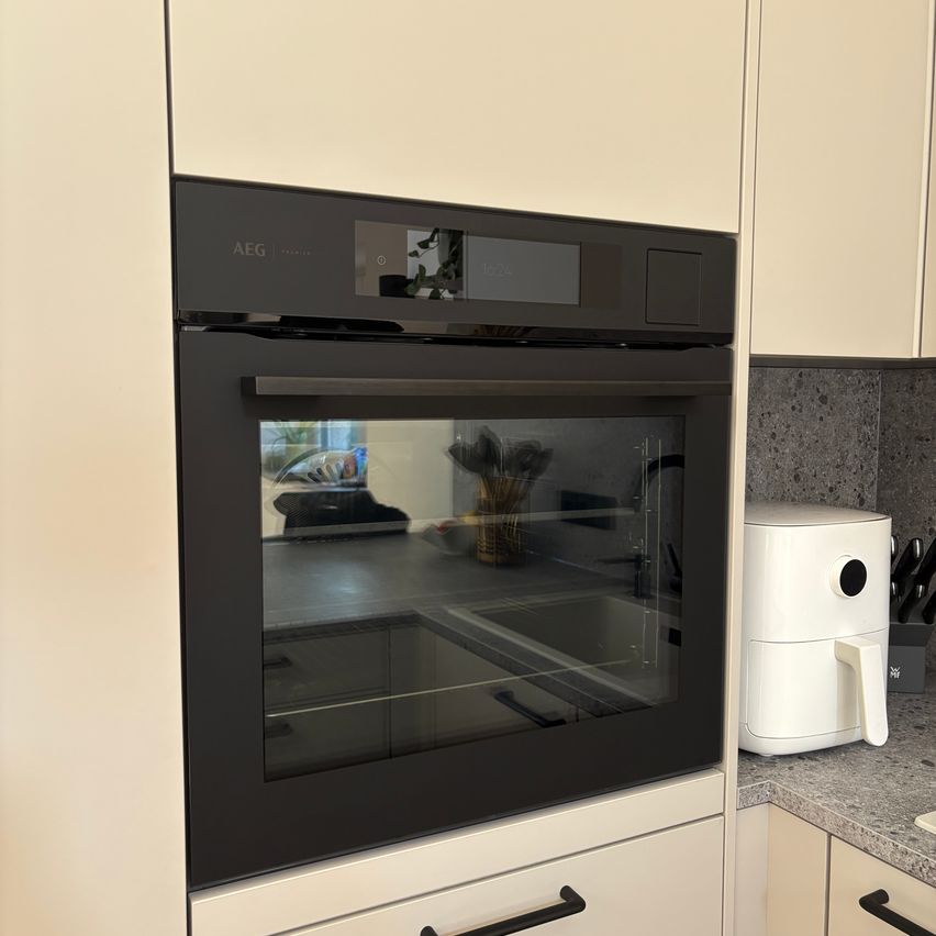 Ein moderner schwarzer AEG-Ofen mit Glastür ist in einer weißen Küche installiert. Daneben steht eine weiße Heißluftfritteuse auf der Arbeitsplatte.