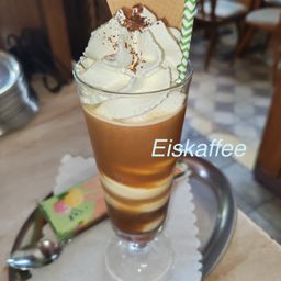 Ein hohes Glas mit einem geschichteten Kaffeegetränk, gekrönt von Schlagsahne und Schokoladenraspeln, mit einem Strohhalm und einem Keks obenauf. Das Getränk ist mit 'Eiskaffee' beschriftet.