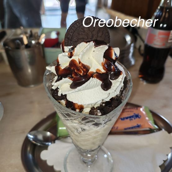 Ein Glas-Sundae mit Eiscreme, Schokoladensauce und einem Oreo-Keks obenauf, mit einer Coca-Cola-Flasche im Hintergrund.