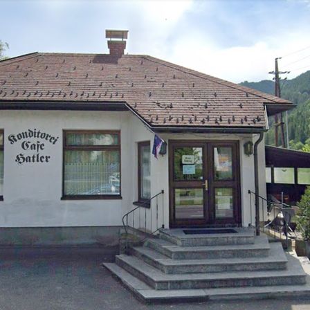 Ein kleines Café mit einem Ziegeldach und einem Schild, das 'Ronditorei Cafe Sattler' steht. Der Eingang ist durch Doppeltüren mit einer Flagge an der Wand.
