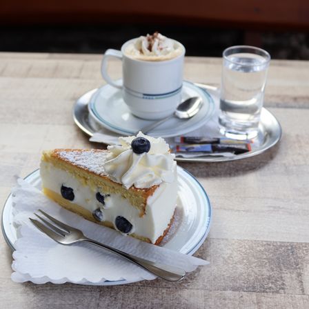 Ein Stück Blaubeer-Käsekuchen mit Schlagsahne und einer Gabel auf einem Teller, begleitet von einer Tasse Kaffee und einem Glas Wasser auf einem Holztisch.