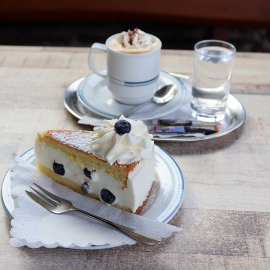 Ein Stück Blaubeer-Käsekuchen mit Schlagsahne und einer Gabel auf einem Teller, begleitet von einer Tasse Kaffee und einem Glas Wasser auf einem Holztisch.