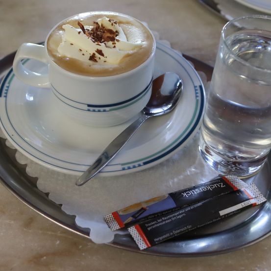 Eine Tasse Kaffee mit Schlagsahne und Schokoladenraspeln auf einer Untertasse, begleitet von einem Löffel und einem Glas Wasser auf einem silbernen Tablett.