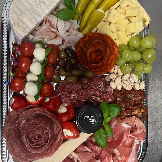 Ein silbernes Tablett zeigt eine Auswahl an Lebensmitteln, darunter geschnittenes Fleisch, Käse, Oliven, Tomaten und Trauben. Ein schwarzer Deckel mit dem Wort 'Gourmet' ist ebenfalls zu sehen.