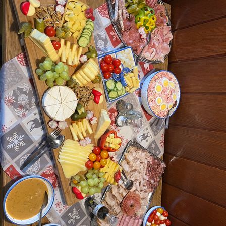Ein Holzbrett mit einer Auswahl an Lebensmitteln wie Käse, Trauben, Tomaten und Fleisch. Es sind auch Schüsseln mit Eiern und eine Schüssel Dip vorhanden.