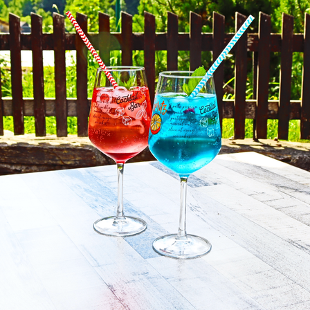 Zwei bunte Cocktails, einer rot und einer blau, stehen auf einem Tisch mit einem Holzzaun im Hintergrund.