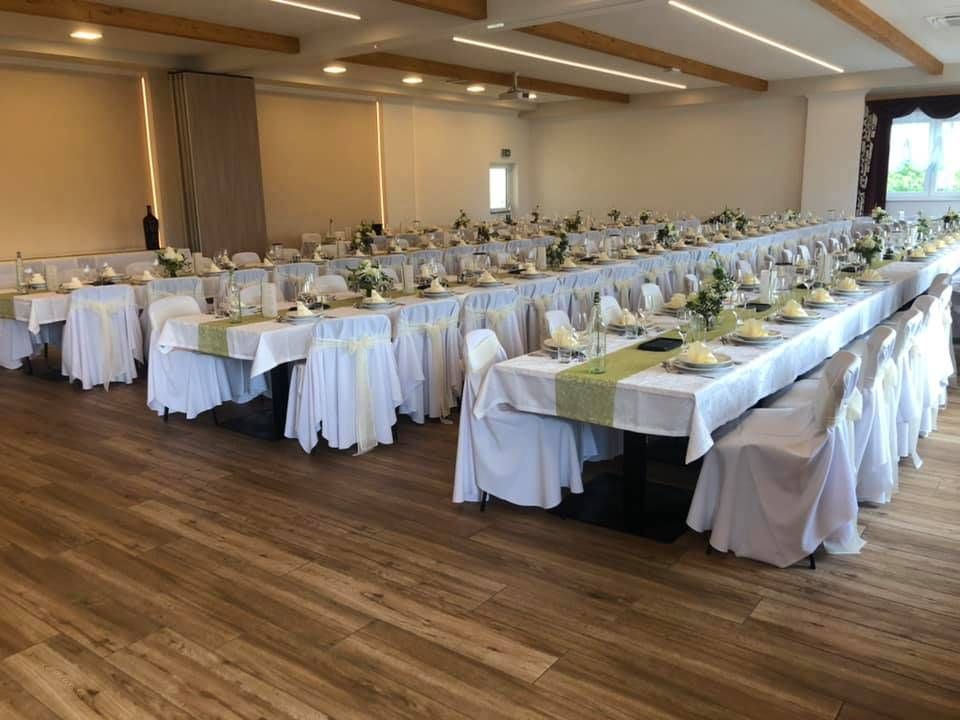 Ein großer Raum mit Holzböden, langen Tischen, die mit weißen Tüchern und grünen Bändern bedeckt sind. Reihen von Stühlen sind mit weißem Stoff bedeckt, und es gibt Blumen auf jedem Tisch.