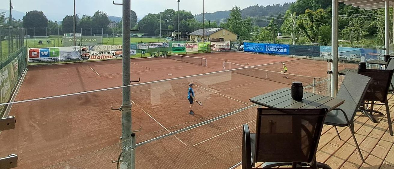 Ein Tennisplatz mit zwei Spielern, einer hält eine Schläger, aus einer Pavillon mit Stühlen und einem Tisch betrachtet. Im Hintergrund ein wolkiger Himmel und entfernte Berge.
