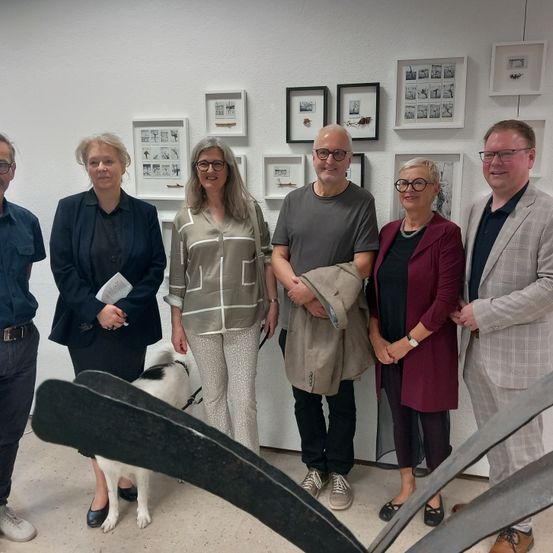 Eine Gruppe von sechs Personen, darunter eine Frau mit einer Leine und einem Hund, steht vor einer Wand mit aufgehängten Bilderrahmen.