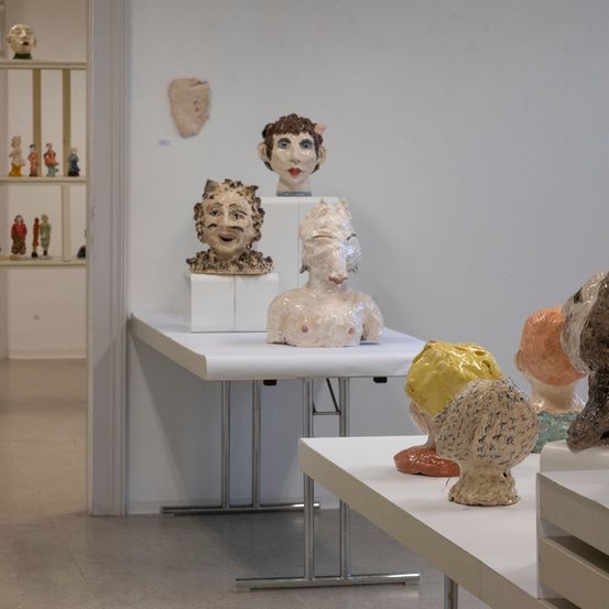 Skulpturen von menschlichen Köpfen sind auf weißen Tischen in einer Galerie ausgestellt. Die Skulpturen bestehen aus verschiedenen Materialien und Farben.