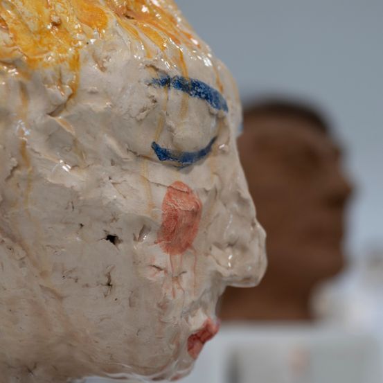 Nahaufnahme einer weißen Keramikskulptur eines Gesichts mit gelber und blauer Farbe und einer Person im Hintergrund
