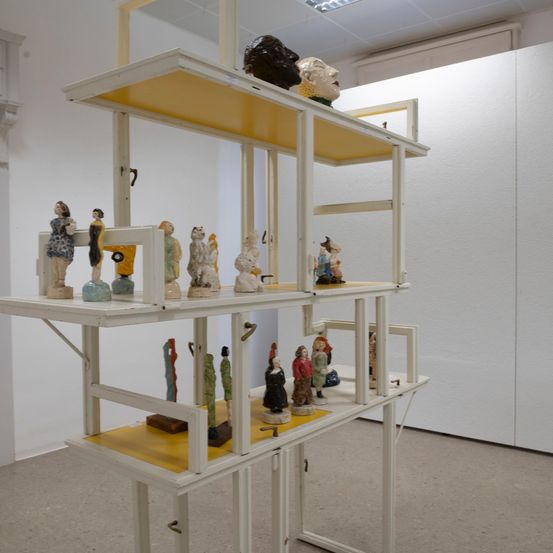 Eine Innenausstellung von weißen Regalen mit gelben Oberseiten. Jedes Regal hat verschiedene Skulpturen, meist von Menschen in verschiedenen Posen. Eine große Skulptur befindet sich auf dem obersten Regal.