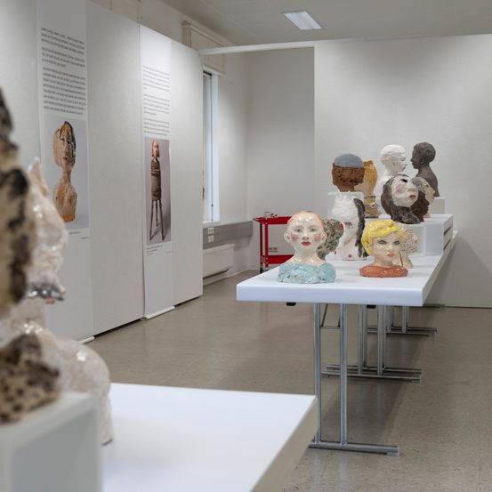 Eine Ausstellung von Skulpturen von Köpfen wird auf weißen Tischen in einem weißen Raum gezeigt.