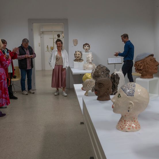 Menschen besuchen eine Indoor-Kunstgalerie mit ausgestellten Kopfbüsten aus Ton, ein Mann hält ein Musikblatt und spricht mit der Gruppe.