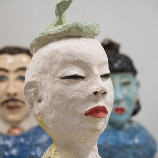 Eine weiße Skulptur einer Frau mit roten Lippen und einem grünen Hut wird vor zwei anderen Skulpturen ausgestellt.