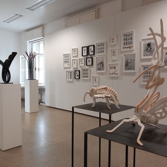 Ein Kunstgalerie mit weißen Wänden, mit Skulpturen und gerahmten Kunstwerken. Die Skulpturen umfassen ein Skelett und abstrakte Stücke auf schwarzen Tischen.