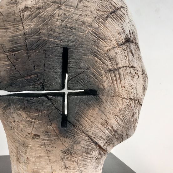 Eine kreuzförmige Skulptur aus einem Baumstamm mit schwarzem Rand und weißem Zentrum wird auf weißem Hintergrund präsentiert.