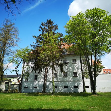 Ein weißes Gebäude mit rotem Dach und vielen Fenstern ist von üppigen grünen Bäumen an einem sonnigen Tag umgeben.
