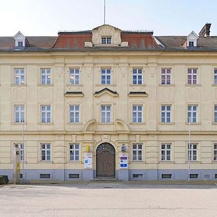 Ein altes architektonisches Gebäude mit vielen Fenstern und einer braunen Tür. Straßenlaternen und Parkschilder befinden sich an den Seiten.
