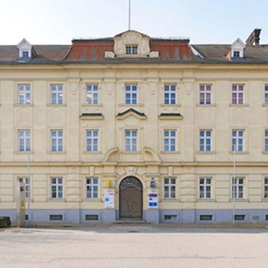 Ein altes architektonisches Gebäude mit vielen Fenstern und einer braunen Tür. Straßenlaternen und Parkschilder befinden sich an den Seiten.