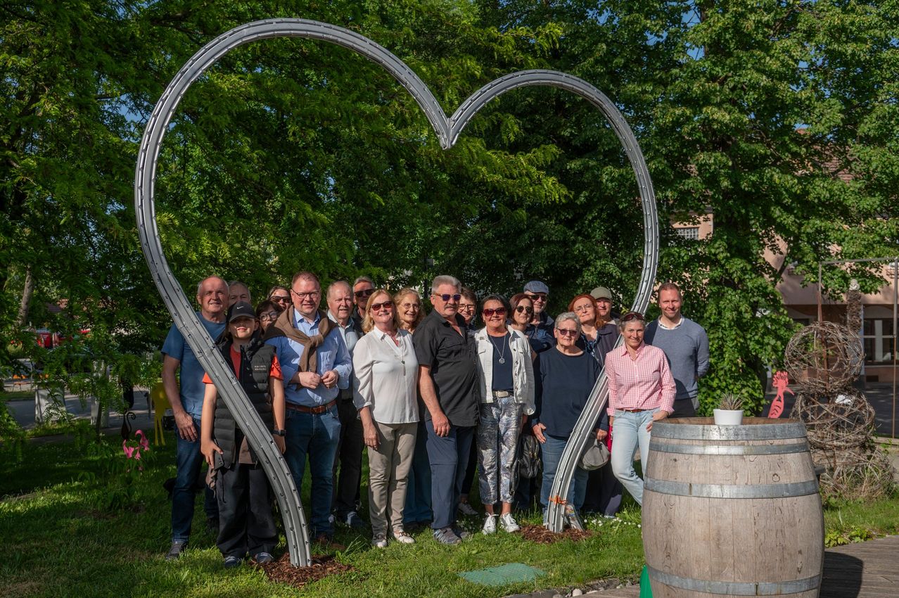Eine Gruppe von Erwachsenen und Kindern posiert für ein Foto vor einer herzförmigen Skulptur in einem Park. Der sonnige Tag verleiht der Szene eine fröhliche Atmosphäre.
