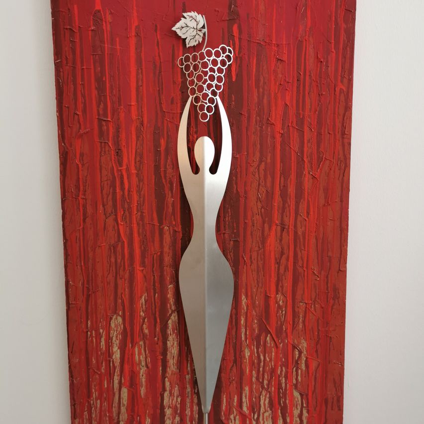 Eine metallische weibliche Figur hält eine Traube in den Händen, vor einem leuchtend roten Hintergrund mit strukturierten Strichen.