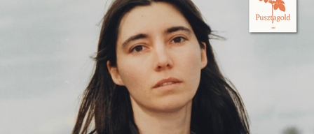 Ein Porträt einer Frau mit langen, dunklen Haaren, die ein hellgraues T-Shirt trägt. Sie hat einen ernsten Gesichtsausdruck und steht vor einem verschwommenen Hintergrund.