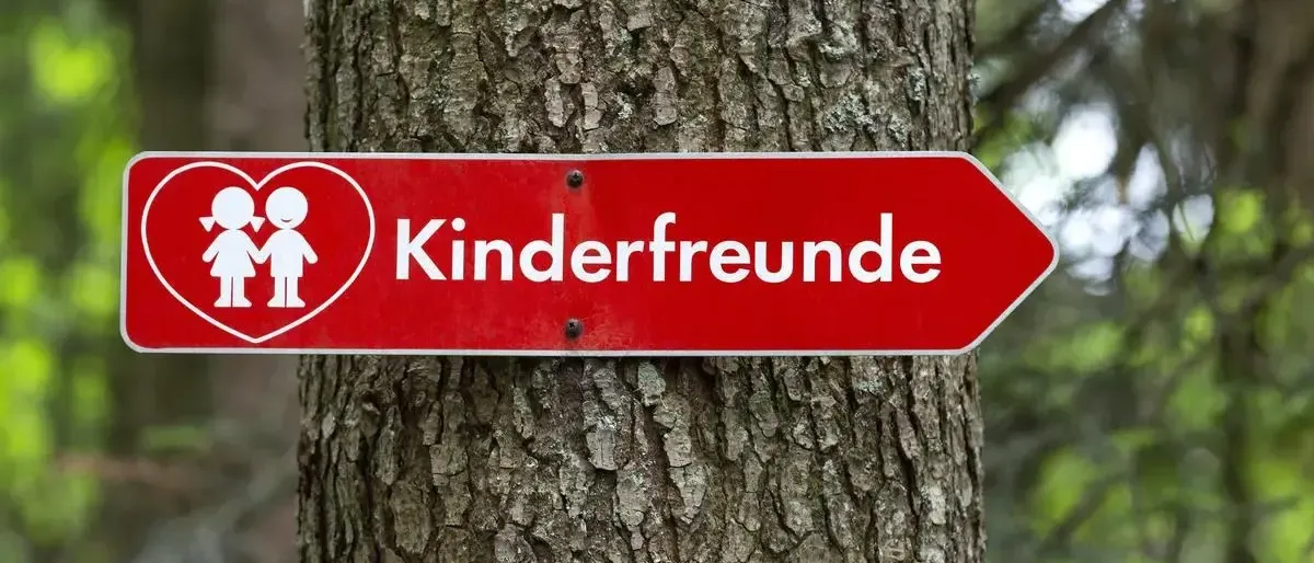 Ein rotes Schild mit dem Text 'Kinderfreunde' ist an einem Baumstamm befestigt. Das Schild hat die Form eines Herzens mit zwei Kindern darin.