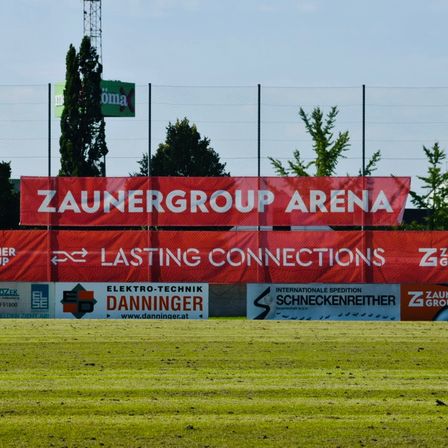 Ein großes rotes Banner mit weißem Text, das 'ZAUNERGROUP ARENA LASTING CONNECTIONS' liest, hängt über einem Feld. Kleinere Banner darunter werben für 'Elektro-Technik Danninger' und 'Internationale Spedition Schneckenreiter.' Grüne Bäume und ein Zaun sind im Hintergrund zu sehen.