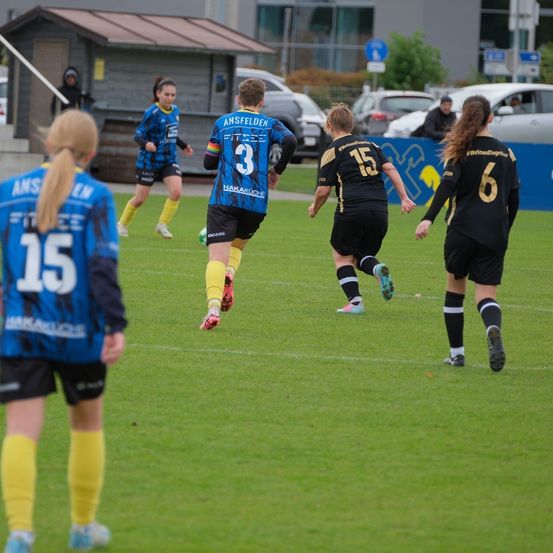 Eine Gruppe Fußballspielerinnen ist auf dem Spielfeld. Eine Spielerin läuft mit dem Ball, während andere verfolgen. Die Spielerinnen tragen nummerierte Trikots. Im Hintergrund befindet sich ein blaues Banner.