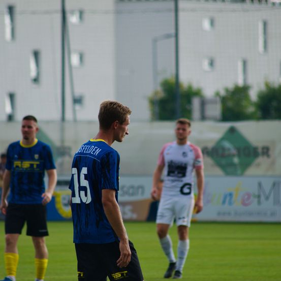 Drei Fußballspieler stehen auf dem Spielfeld, einer von ihnen trägt ein blaues Trikot mit der Nummer '15'. Der Spieler auf der rechten Seite trägt ein weißes Trikot mit einem Logo. Im Hintergrund befindet sich ein Banner mit Text und Logos.