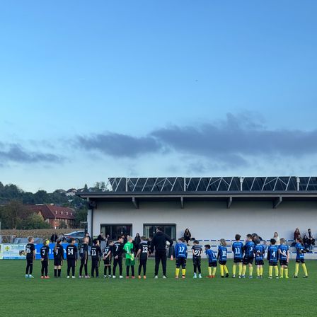 Eine Gruppe junger Fußballer in blauen und gelben Trikots steht auf einem Spielfeld vor einem Gebäude. Zuschauer stehen dahinter und beobachten das Spiel.