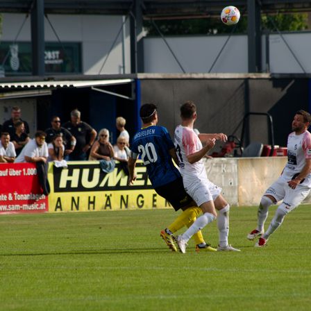 Vier Fußballspieler sind auf dem Platz, einer in Blau springt, um den Ball zu köpfen. Zuschauer beobachten hinter einer Mauer mit einem Banner.