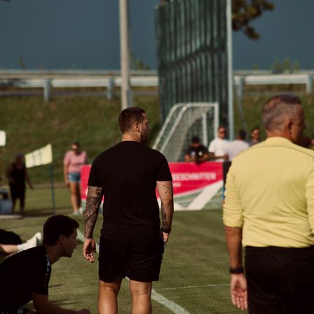 Mehrere Personen befinden sich auf einem Fußballfeld; eine kniet, eine andere mit Tattoos steht, und zwei Schiedsrichter sind anwesend. Im Hintergrund steht ein Tor.